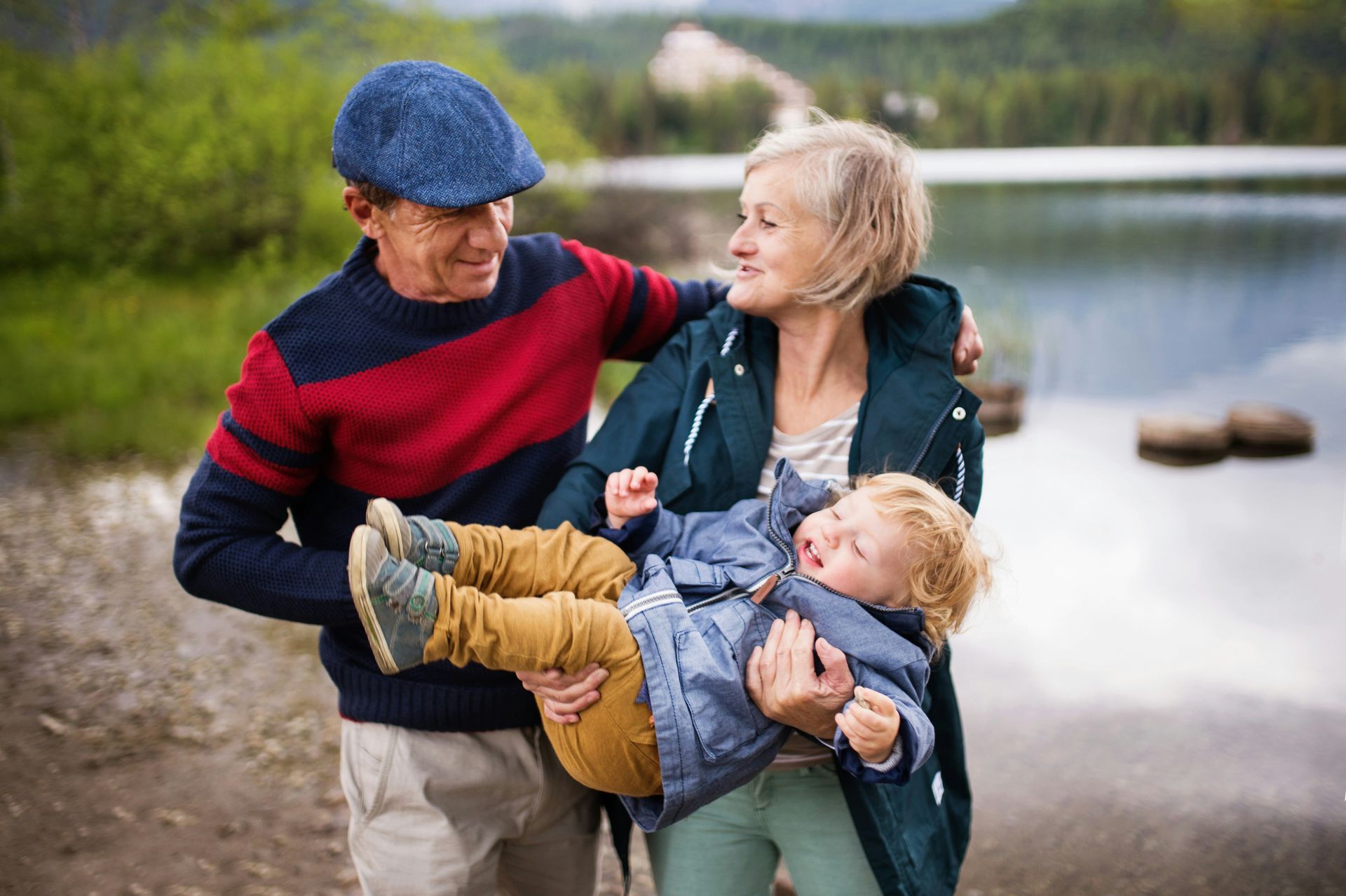 Smiling couple outdoors by a lake, holding a bundled child and wearing autumn clothes