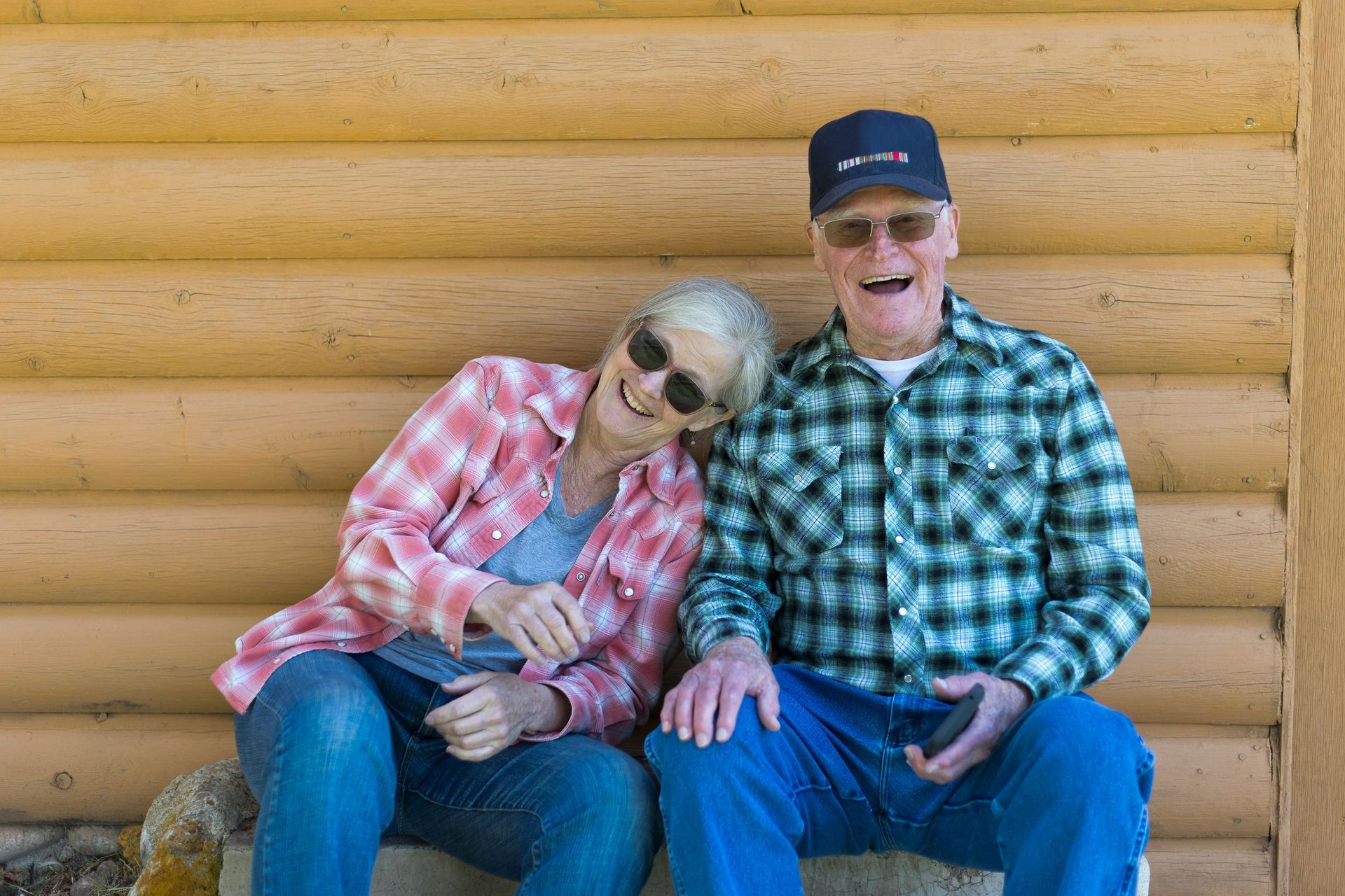 Two smiling people sitting together on a bench in front of a log wall, one leaning on the other