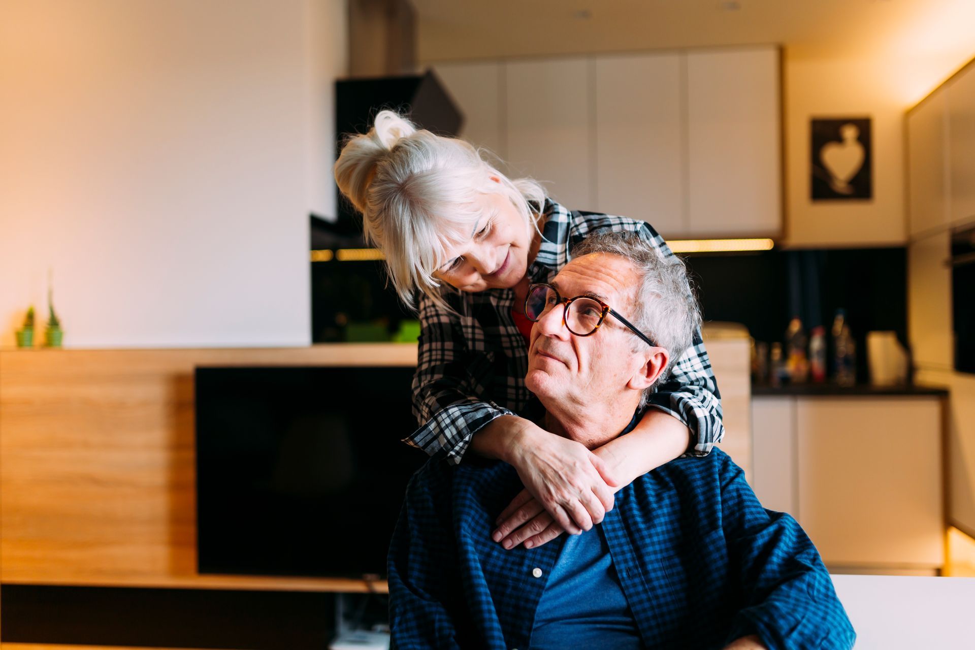A smiling woman hugs an older man from behind in a warm living room.