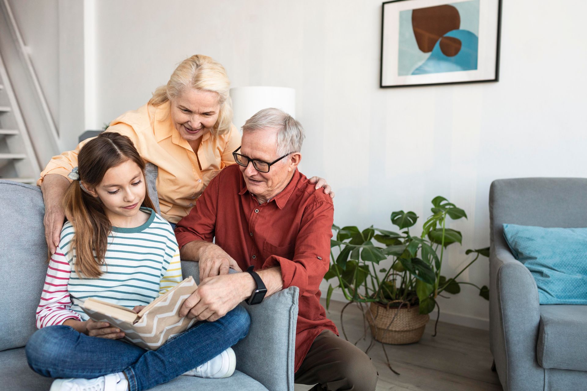 Three people sit on a couch reading a book in a bright living room with plants and a gray chair.