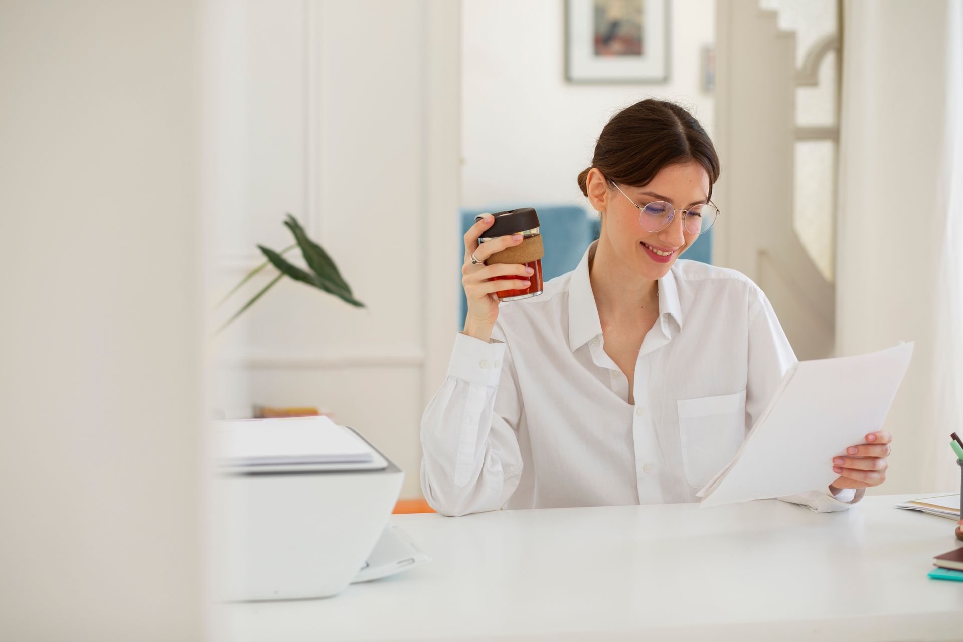 Woman in a white shirt smiling at a desk, holding a drink and reading a paper in a bright office