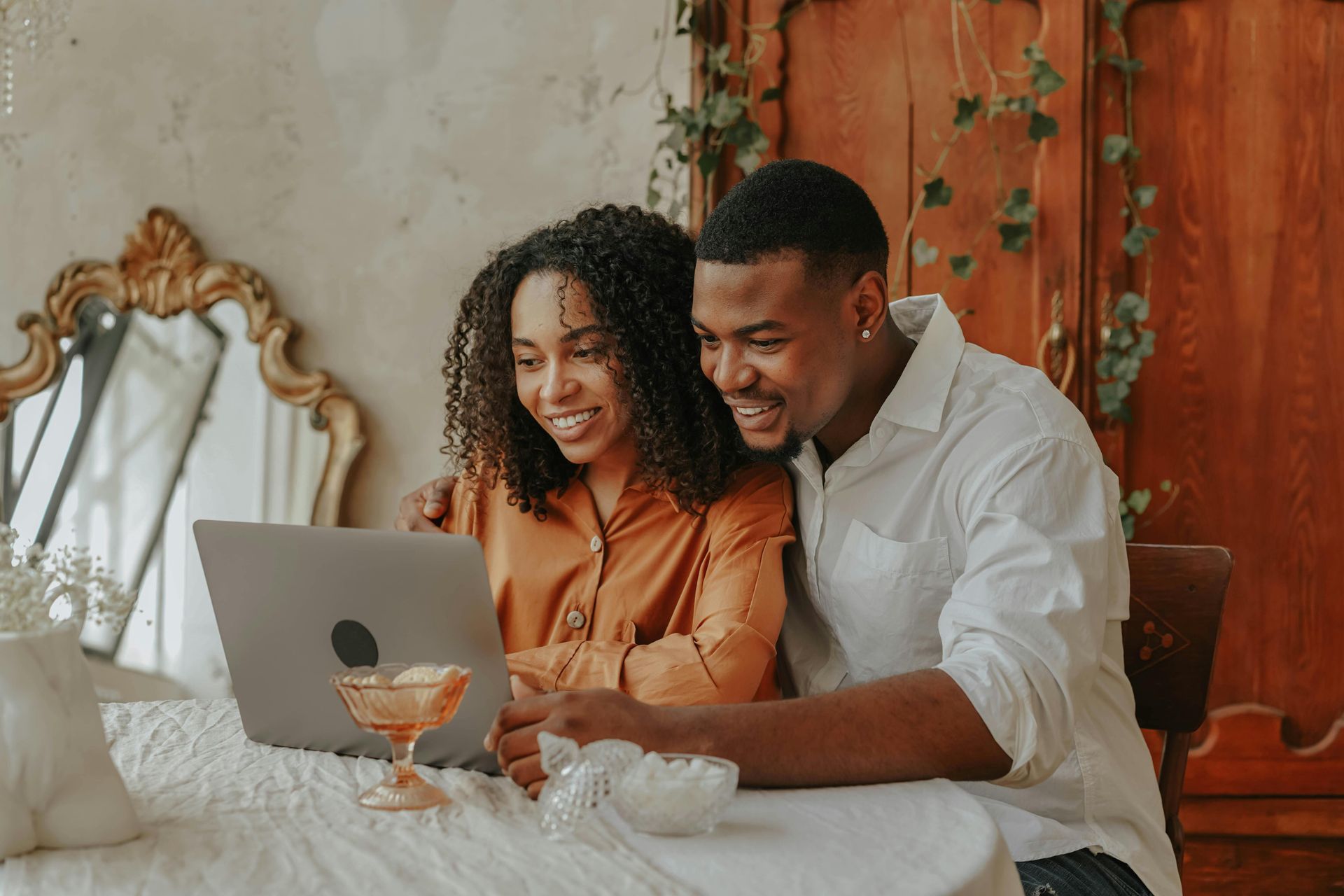 Two people smiling at a laptop in a warm, cozy dining room with a table and ornate mirror.