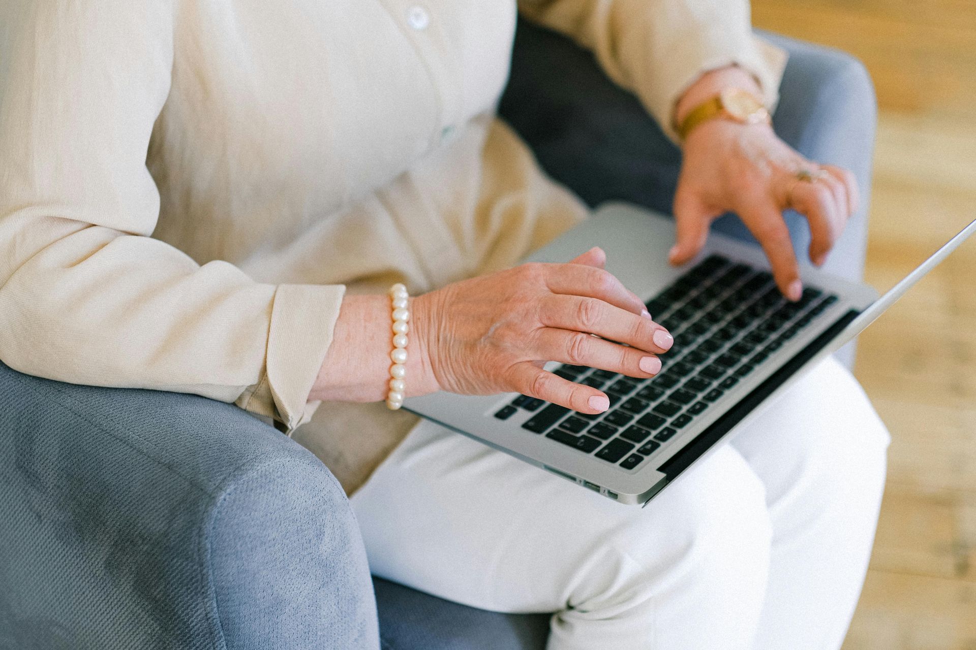 Person typing on a laptop while seated on a couch, wearing a cream sweater and white pants