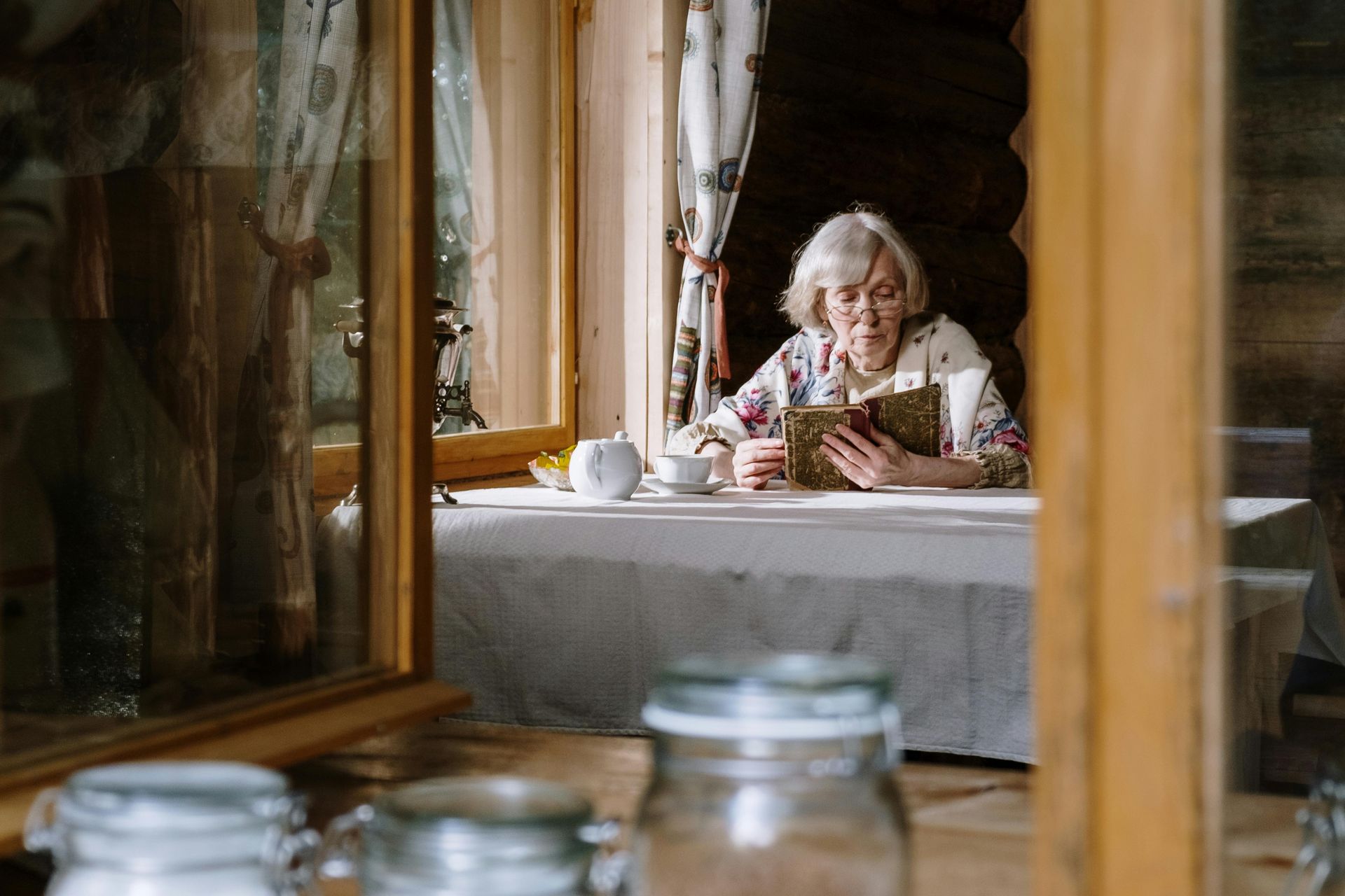 Woman seated at a table in a rustic café, looking down at a cup, framed by wooden window reflections