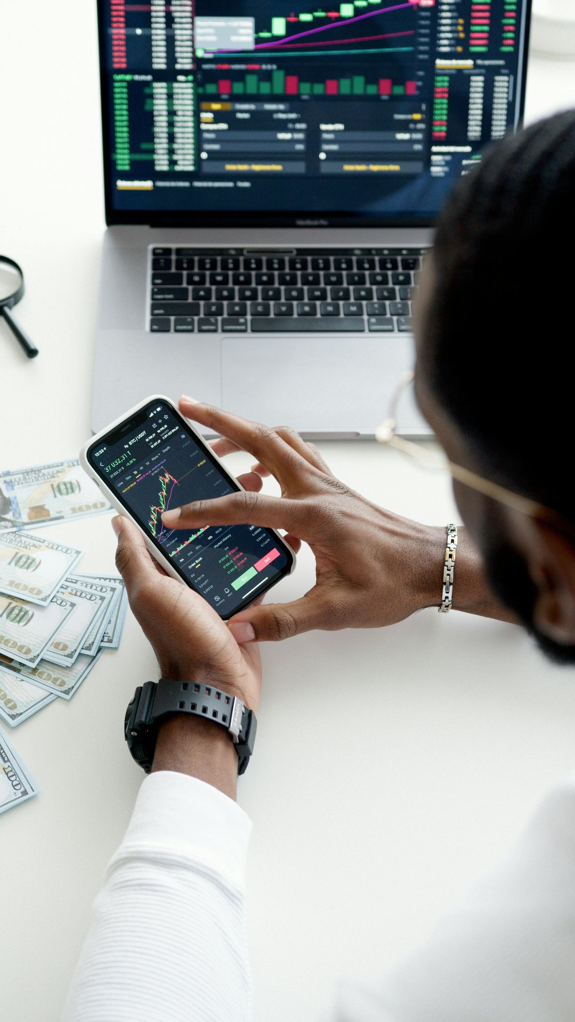 Person checking stock charts on a smartphone at a desk with a laptop displaying trading data.