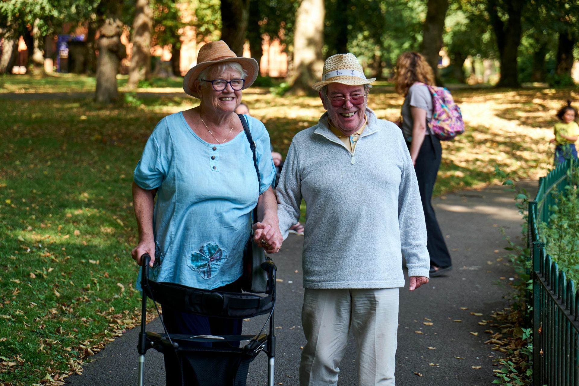 Two people walking arm in arm on a tree-lined park path, one using a rollator walker.