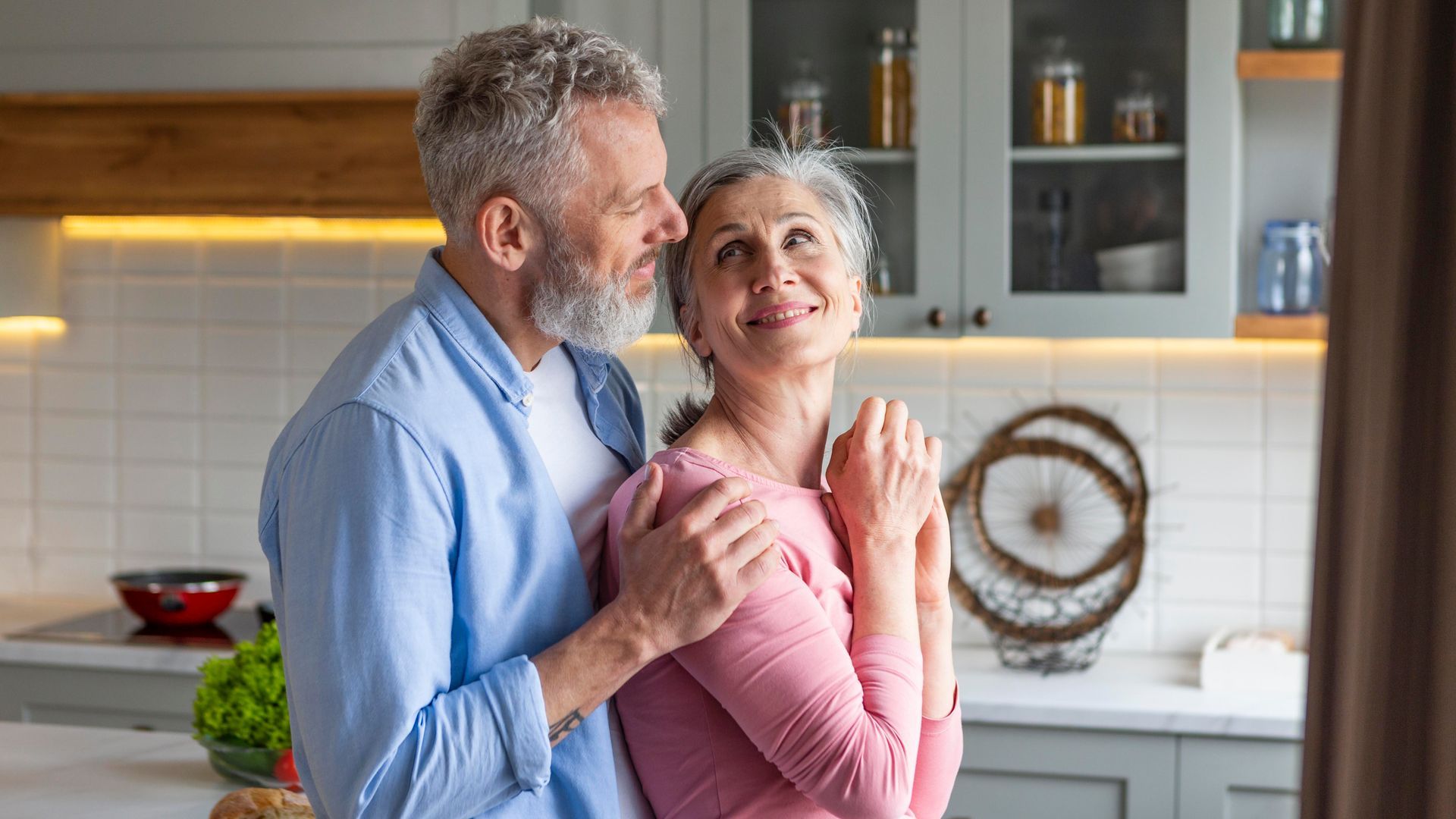 Smiling couple in a bright kitchen, standing close and holding hands near the counter.