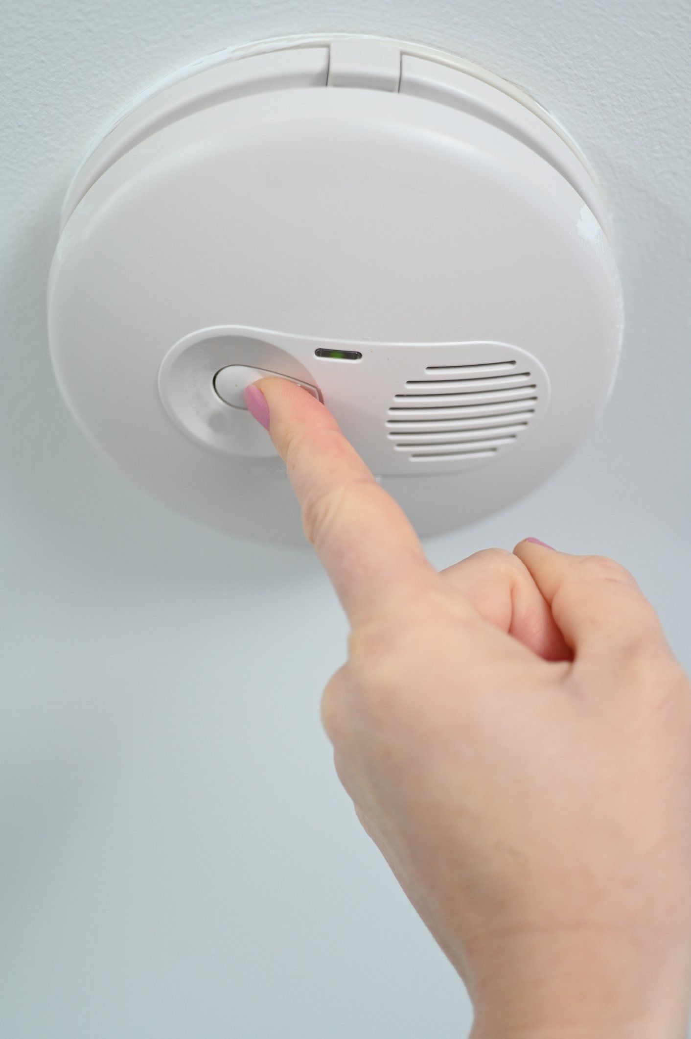 A hand presses the test button on a round white smoke alarm mounted on a ceiling.