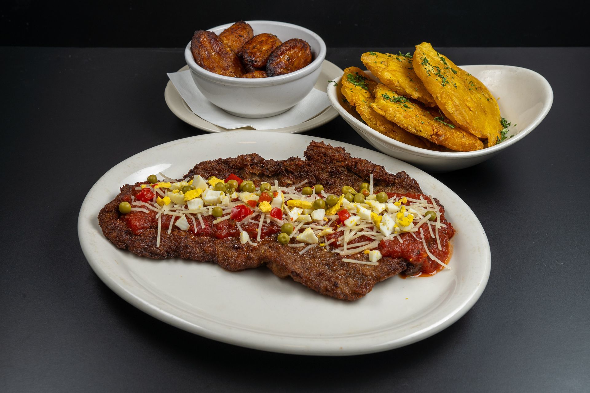 Grilled steak topped with onions and herbs on a plate, with a bowl of fried plantains and tostones