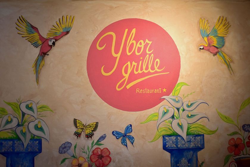 Colorful mural with “Ybor grille” logo, birds, butterflies, flowers, and plants on a cream wall