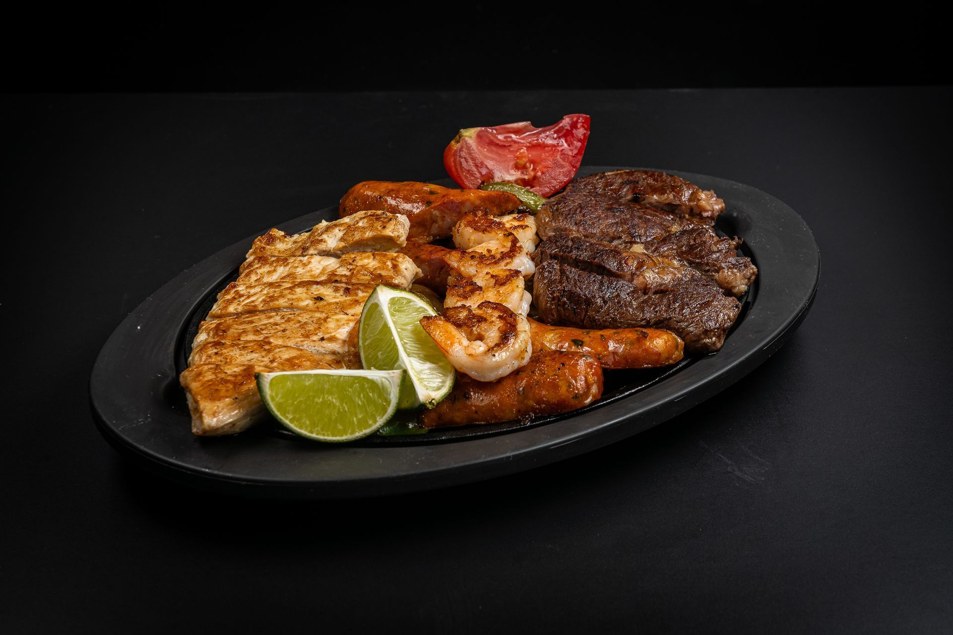 Grilled meat platter with chicken, steak, sausage, lime wedges, and a red pepper on a black plate