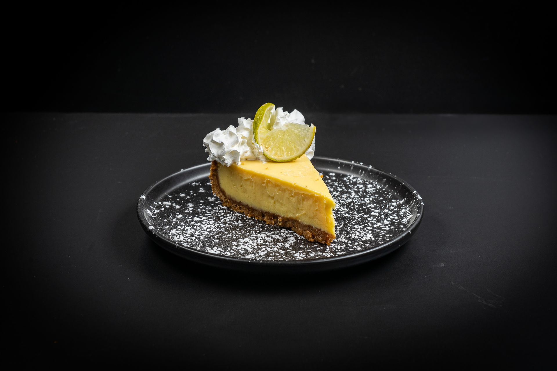 Slice of lemon cheesecake on a black plate, topped with whipped cream and a lemon wedge on a dark background