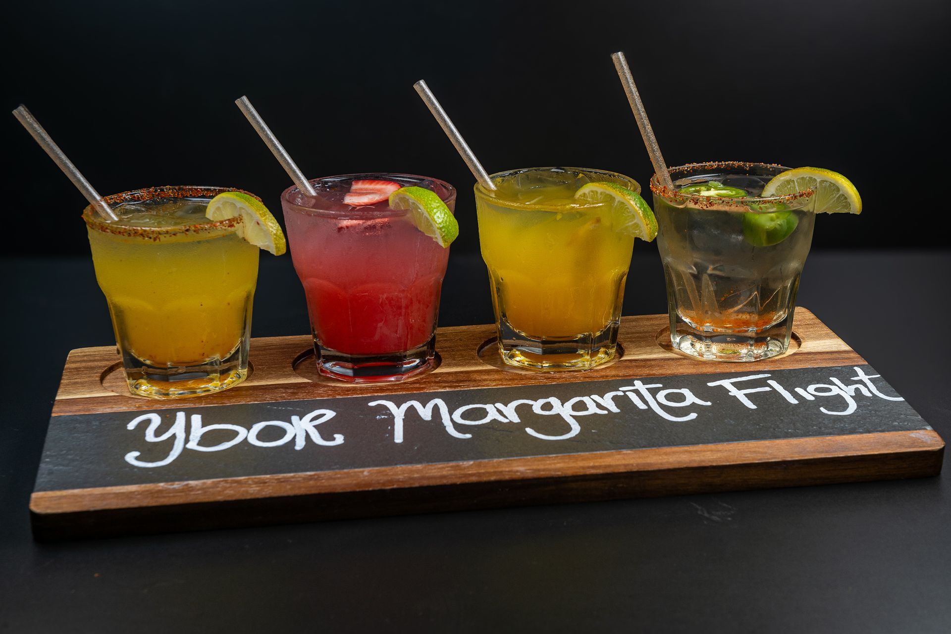 Four colorful margarita shots with lime wedges on a wooden board labeled “Ybor Margarita Flight”