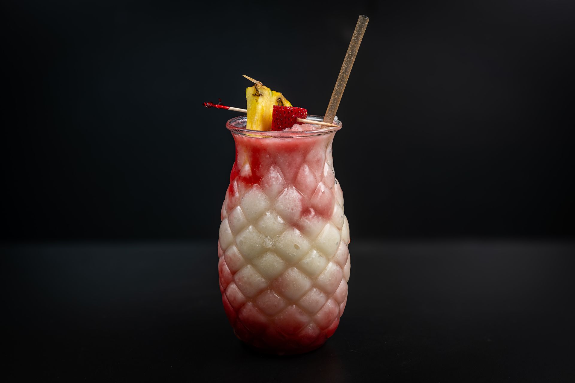 Pink layered cocktail in a textured glass, garnished with pineapple and cherry on a dark background