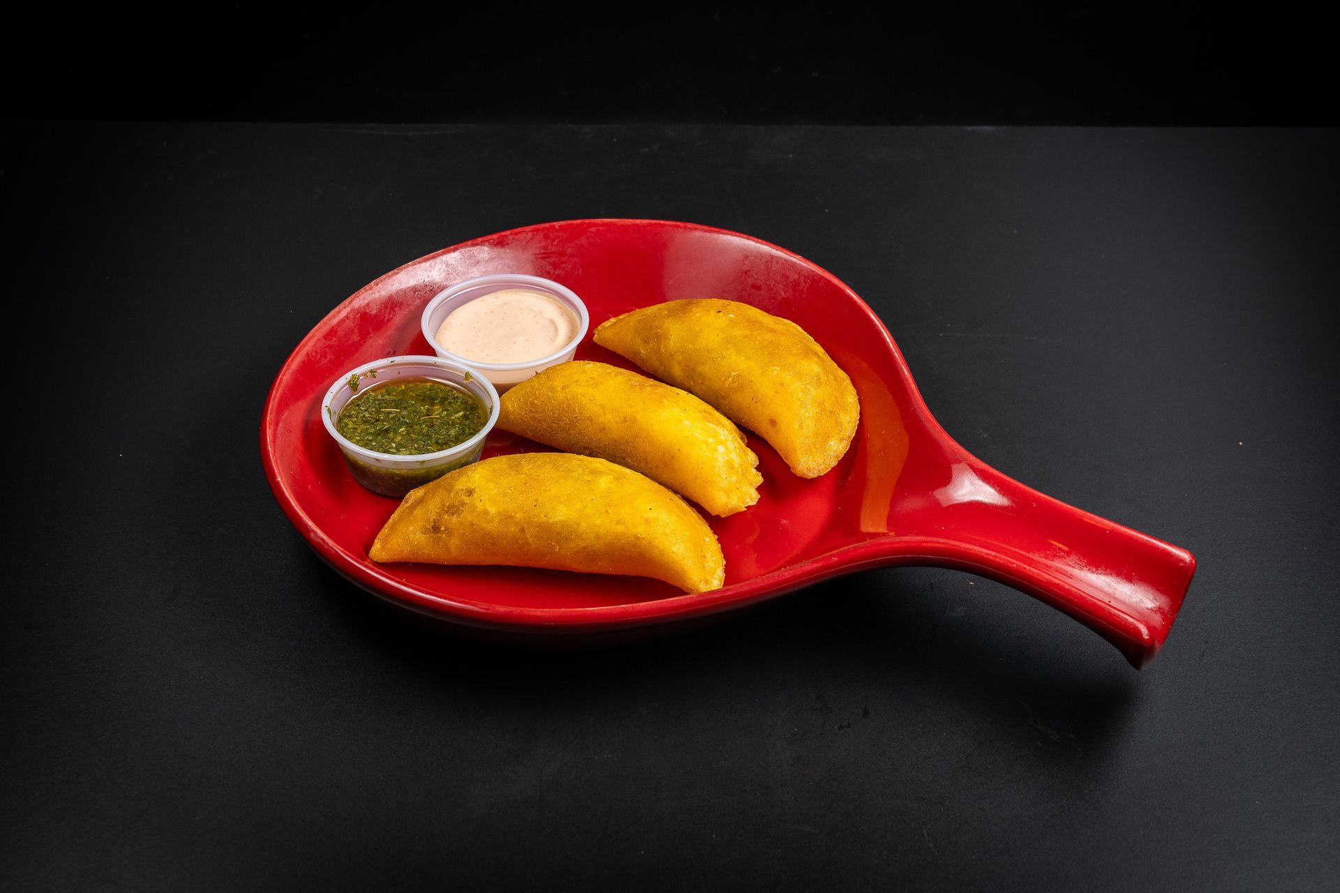 Red pan with fried empanadas and two dipping sauces on a black background