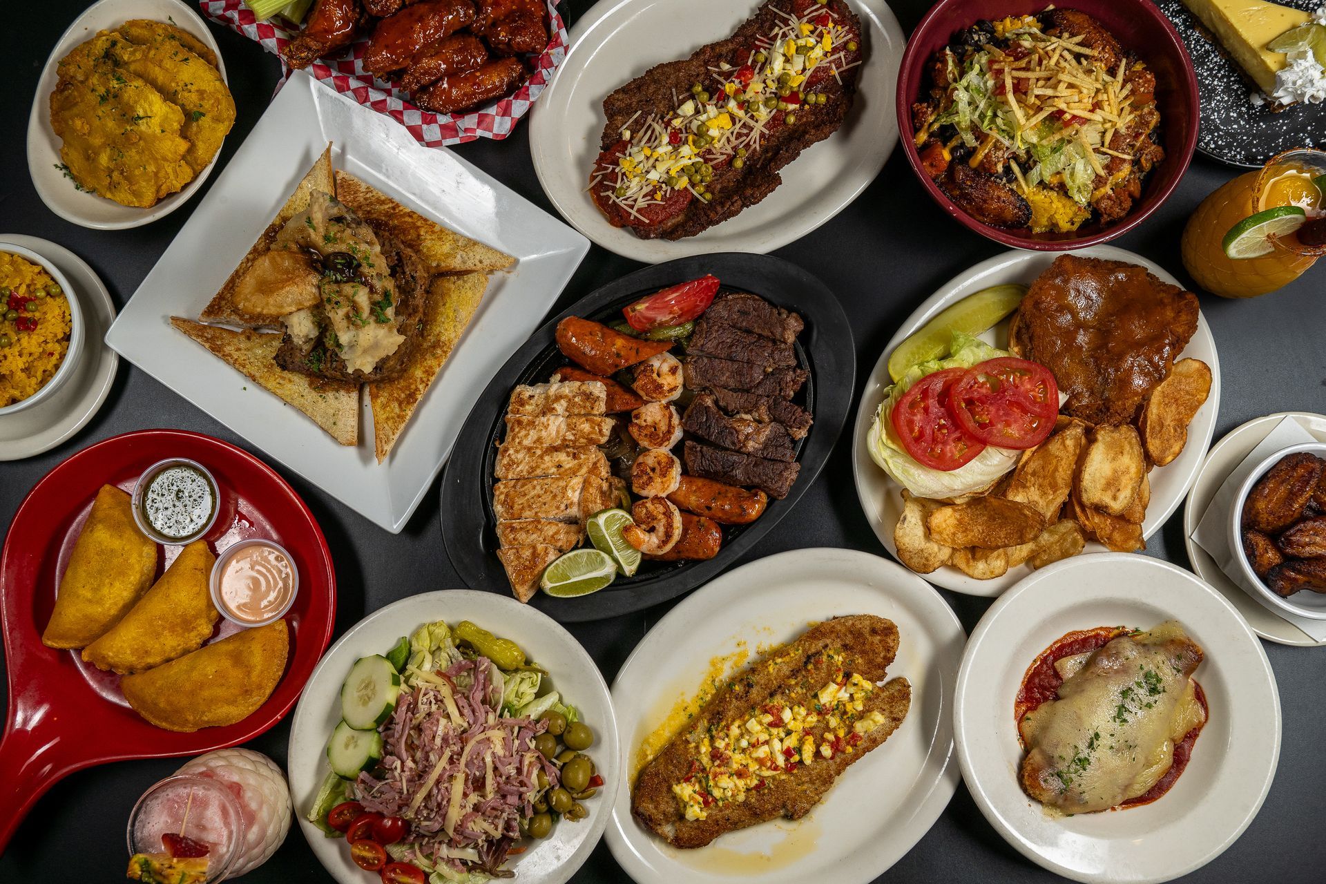 Assorted plated dishes and grilled meats arranged on a dark table in a colorful overhead food spread