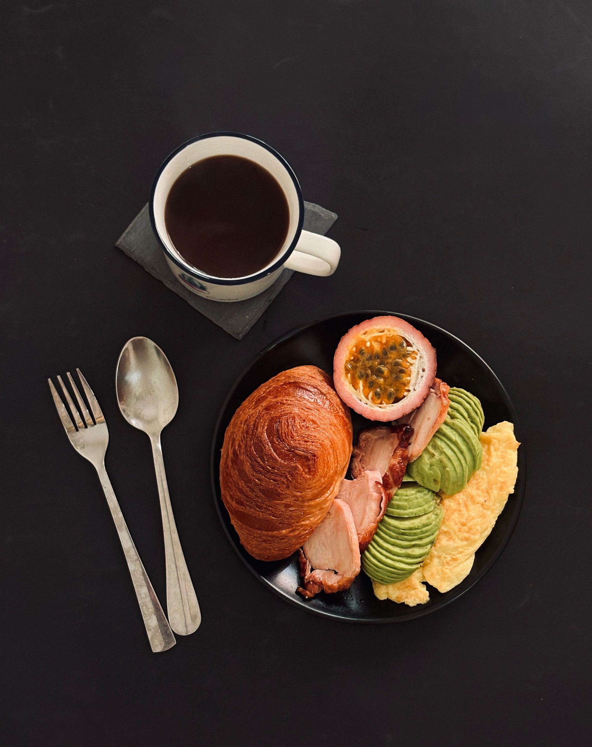 Breakfast plate with croissant, avocado toast, eggs, bacon, yogurt, and a cup of coffee on a dark table