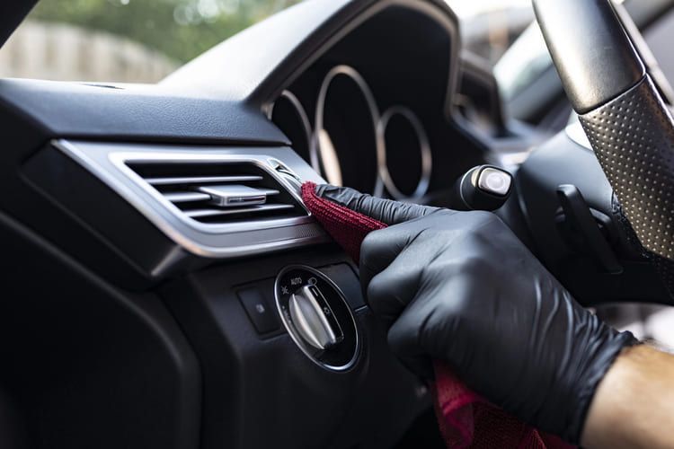 A person wearing black gloves is cleaning the dashboard of a car with a microfibre cloth.