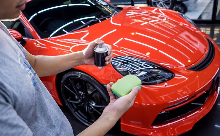 A man is polishing a red sports car with a sponge and spray bottle.