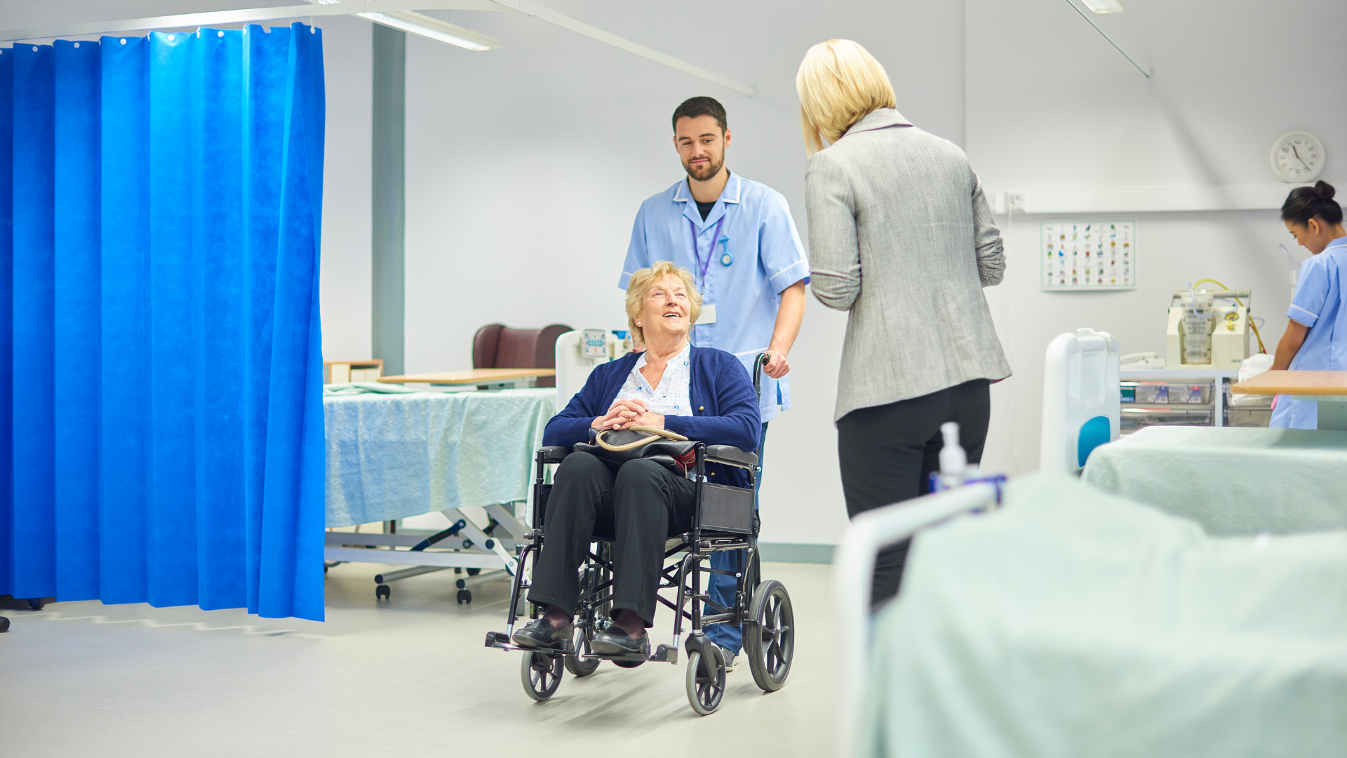 Hospital room with a patient in a wheelchair, a clinician nearby, and another person standing beside the bed