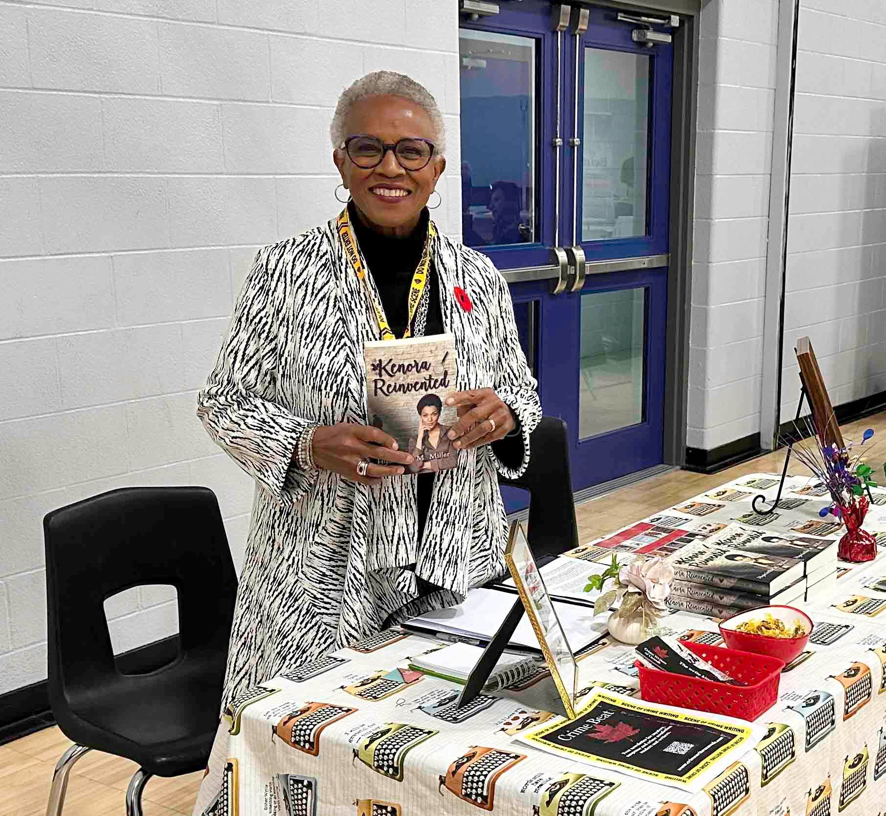 Woman holding a book at a table, smiling. She wears a black and white jacket. Books and items are on display.
