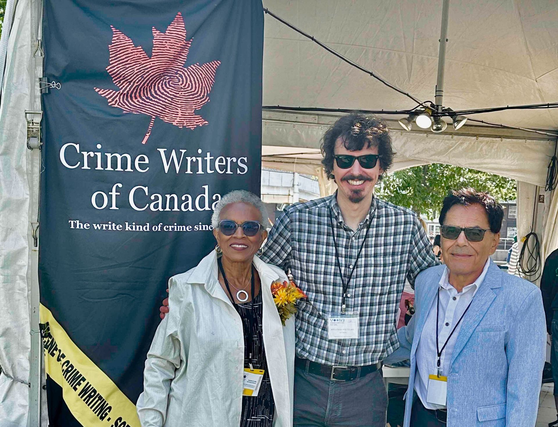 Three people stand in front of a Crime Writers of Canada banner at an outdoor event.
