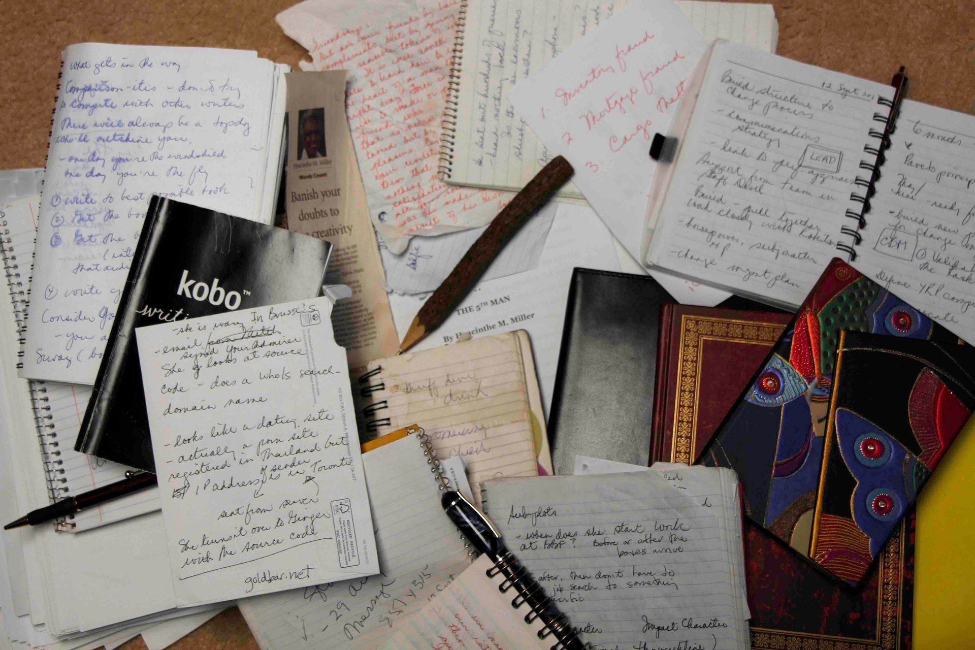 Pile of notebooks and papers; handwritten notes, a Kobo reader, and pens are scattered.