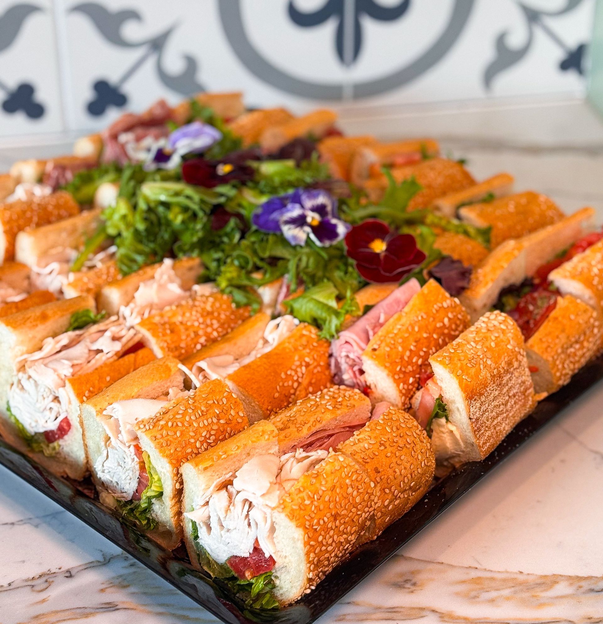 Tray of sliced sandwiches, garnished with greens and edible flowers, on a decorative tile background.