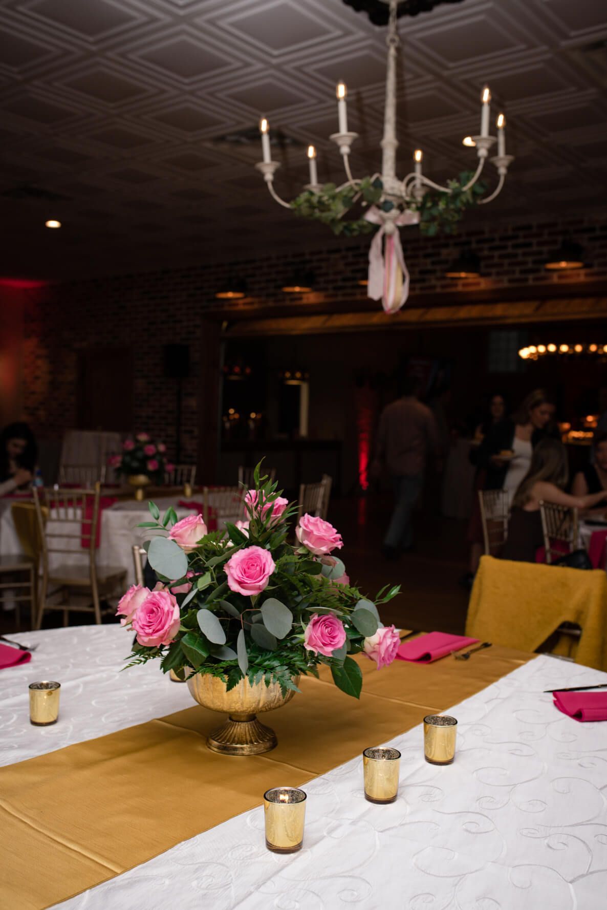 Elegant table setting with pink roses, gold accents, and a chandelier in a banquet hall.
