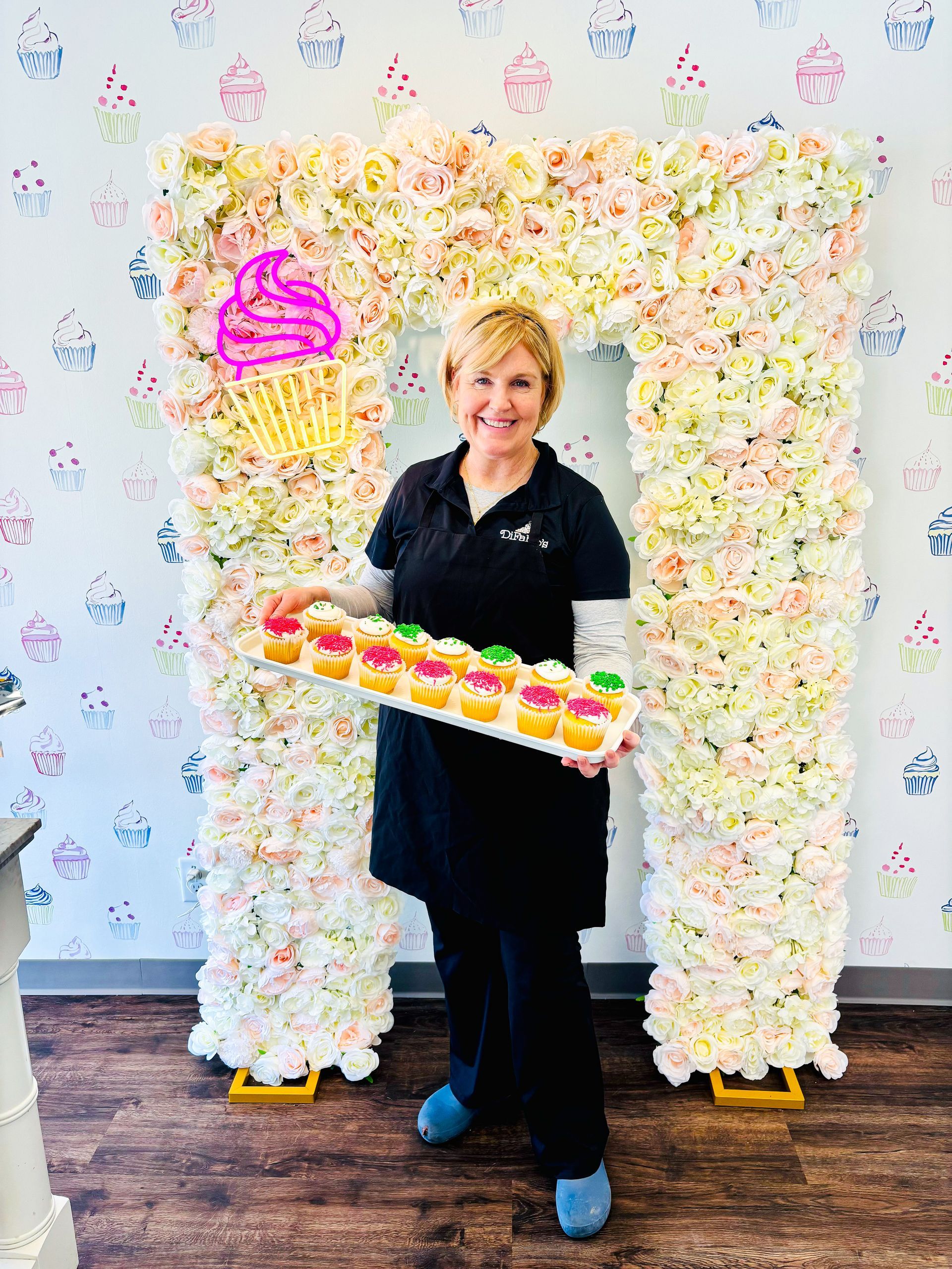 Woman in black holding a tray of colorful cupcakes in front of a floral backdrop and neon pink sign