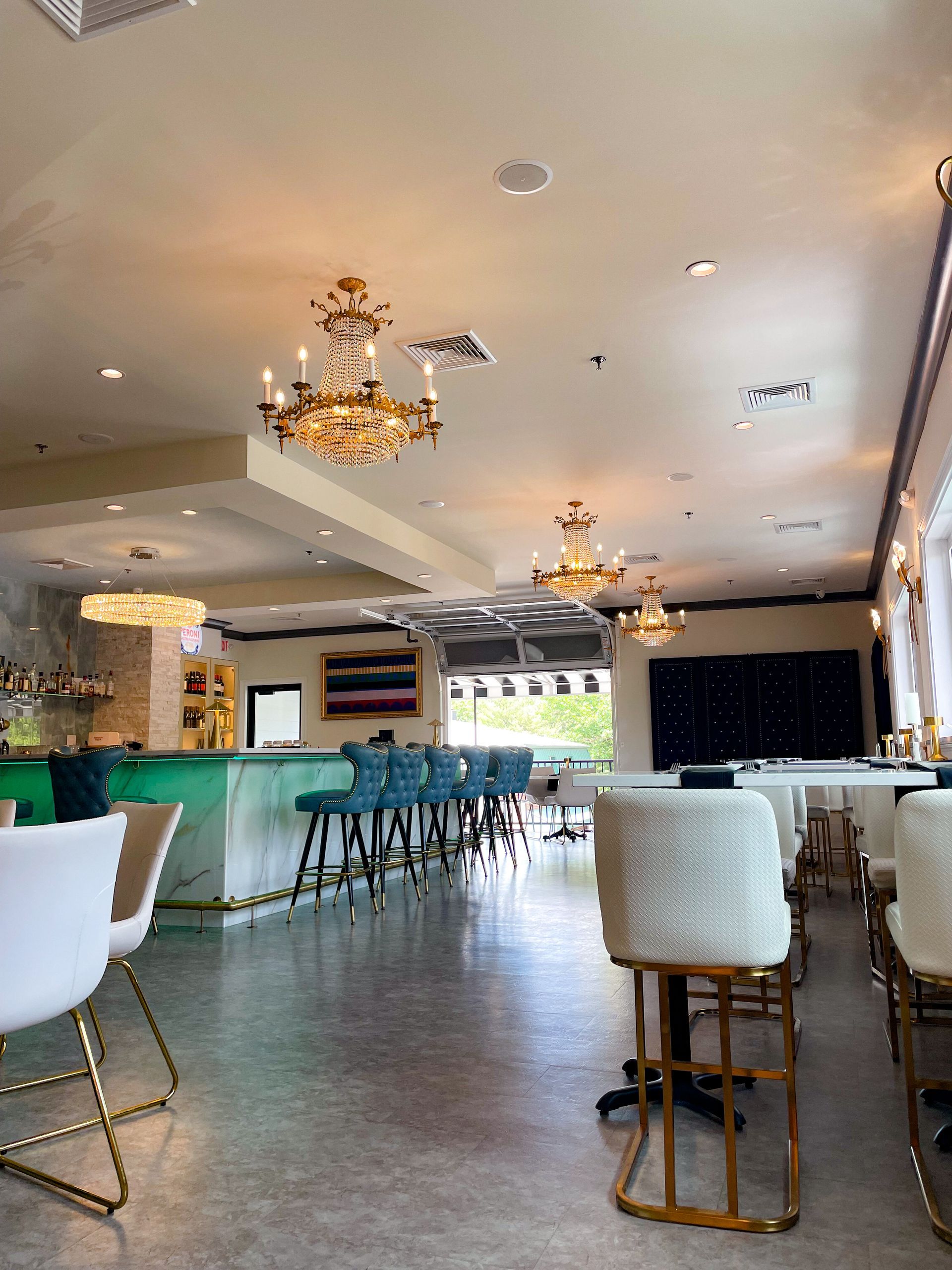 Interior of a restaurant with ornate chandeliers, a turquoise bar, and white chairs with gold accents.