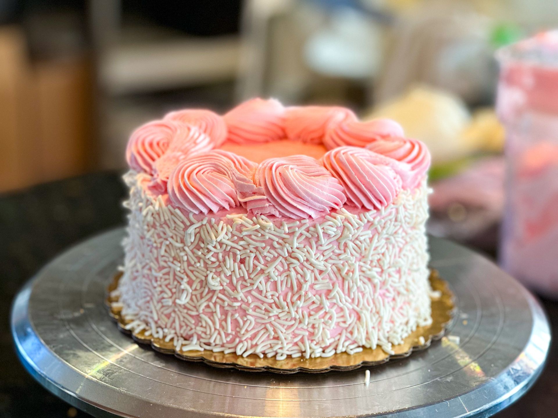 Pink frosted cake with white sprinkles and swirls, on a silver turntable.