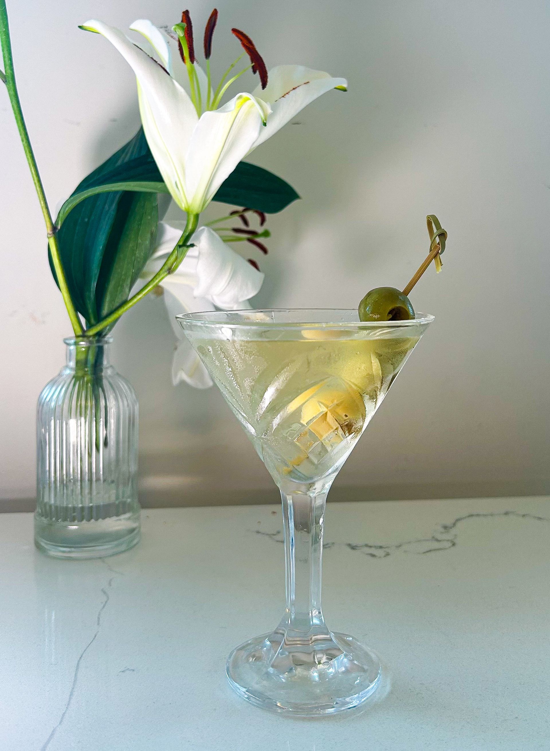 Martini in a stemmed glass with olives, next to a vase with white lilies.