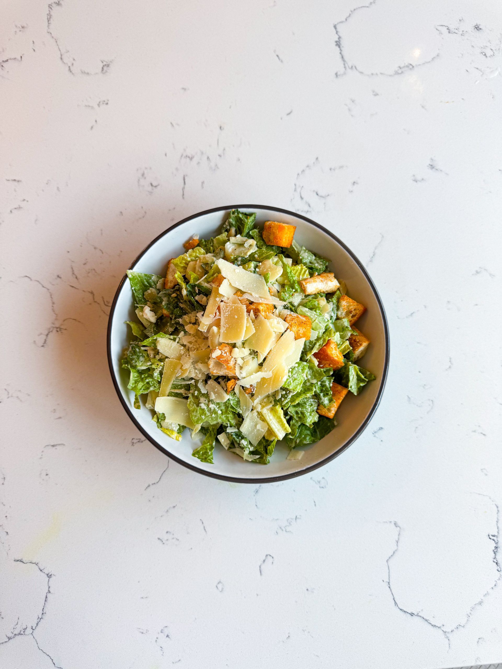 Caesar salad in a white bowl, with parmesan and croutons, on a marble surface.