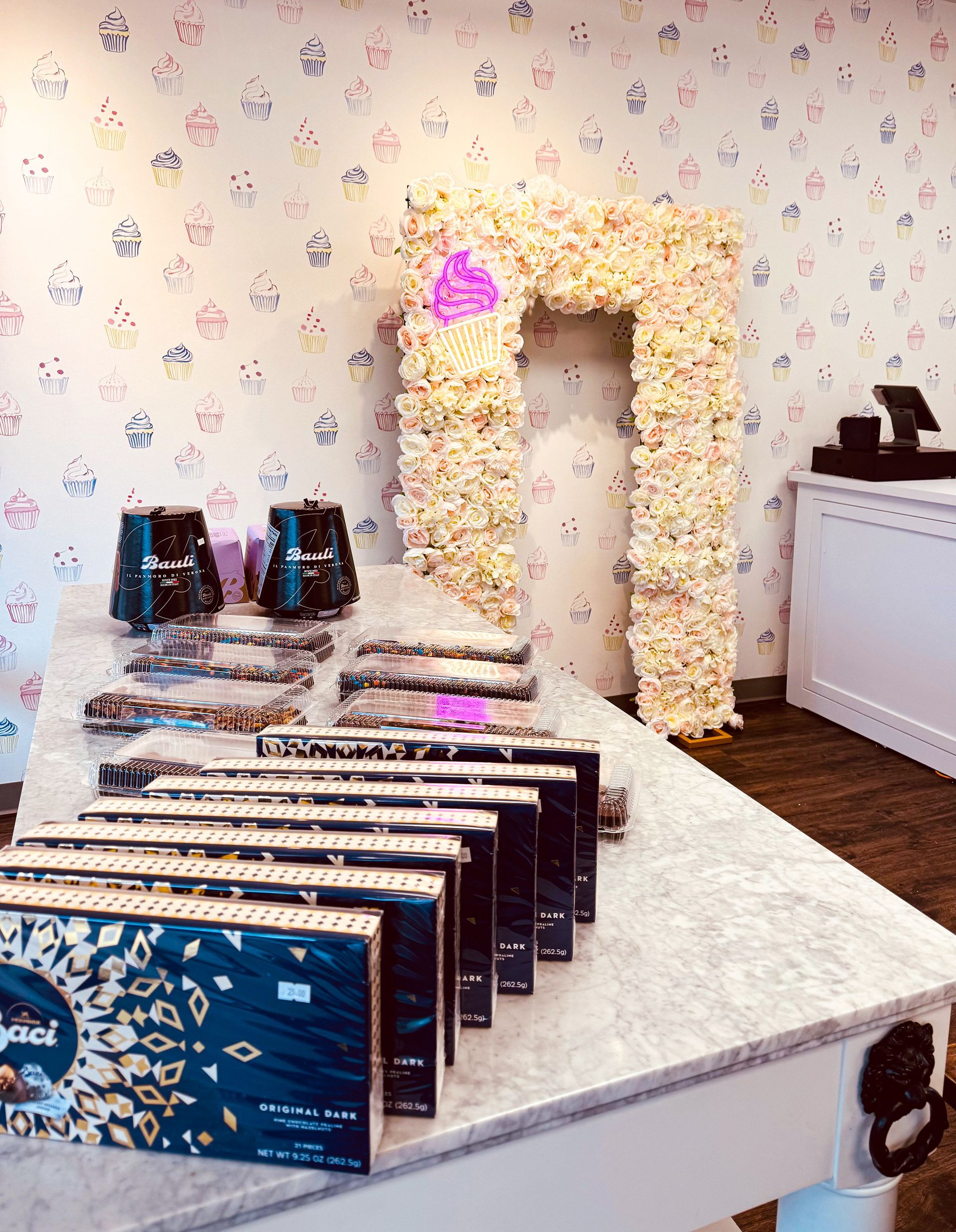 White dessert table with chocolate bars and cups, in front of a flower-covered archway at an event.