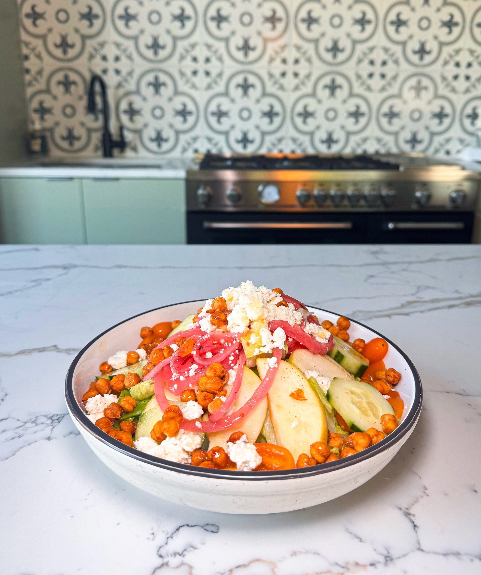 Salad in bowl on marble counter in kitchen, with chickpeas, pickled onion, cheese, cucumbers, and apples.