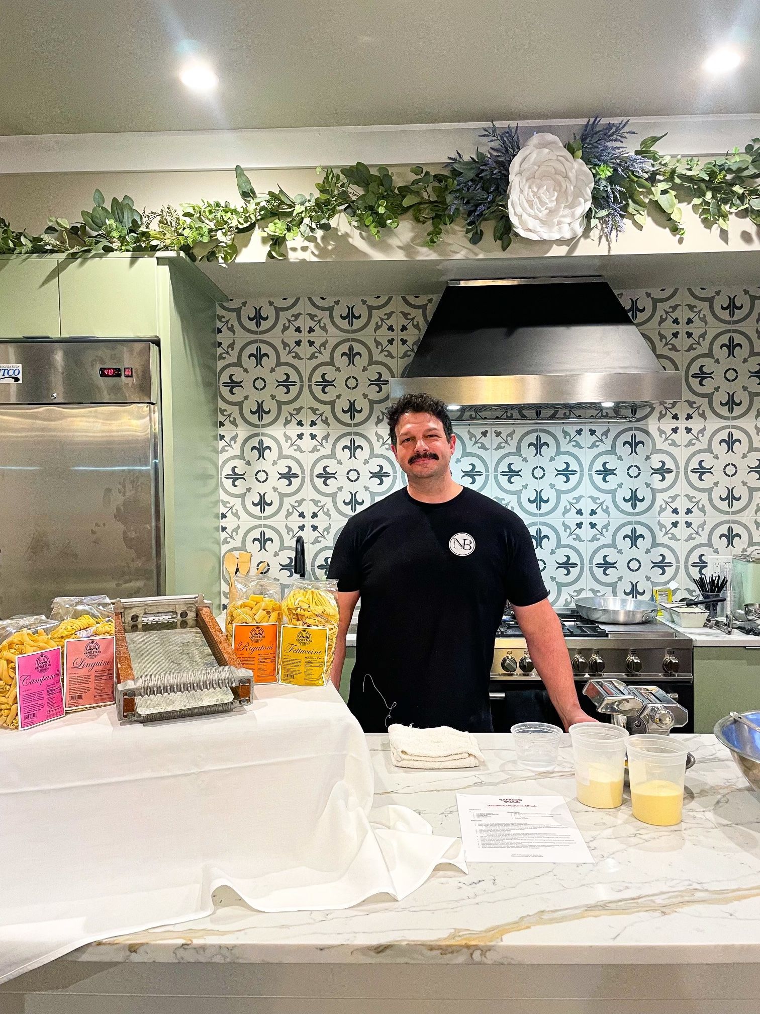Three people standing behind a kitchen island at a buffet, with food trays and hanging greenery overhead