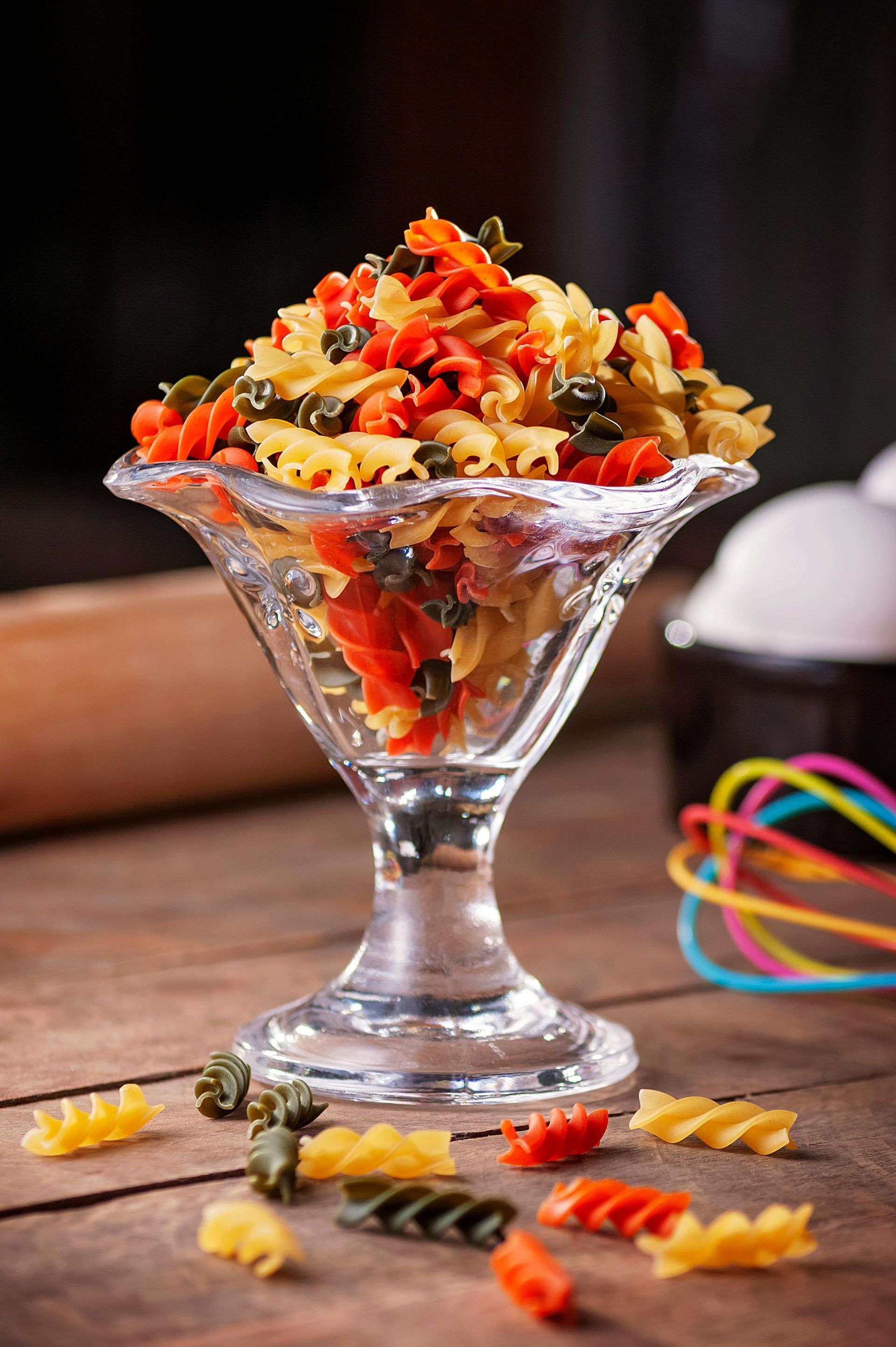 Glass dessert dish filled with colorful pasta on a wooden table, with pasta scattered around it