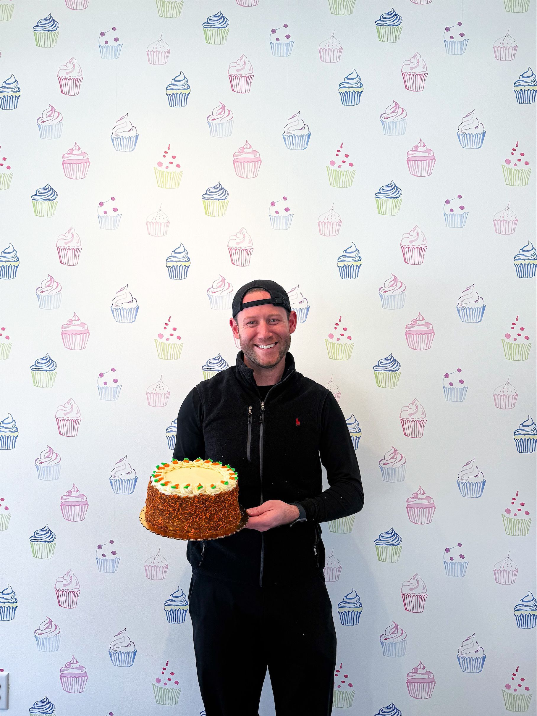 Person holding a large cake and smiling in front of patterned wallpaper