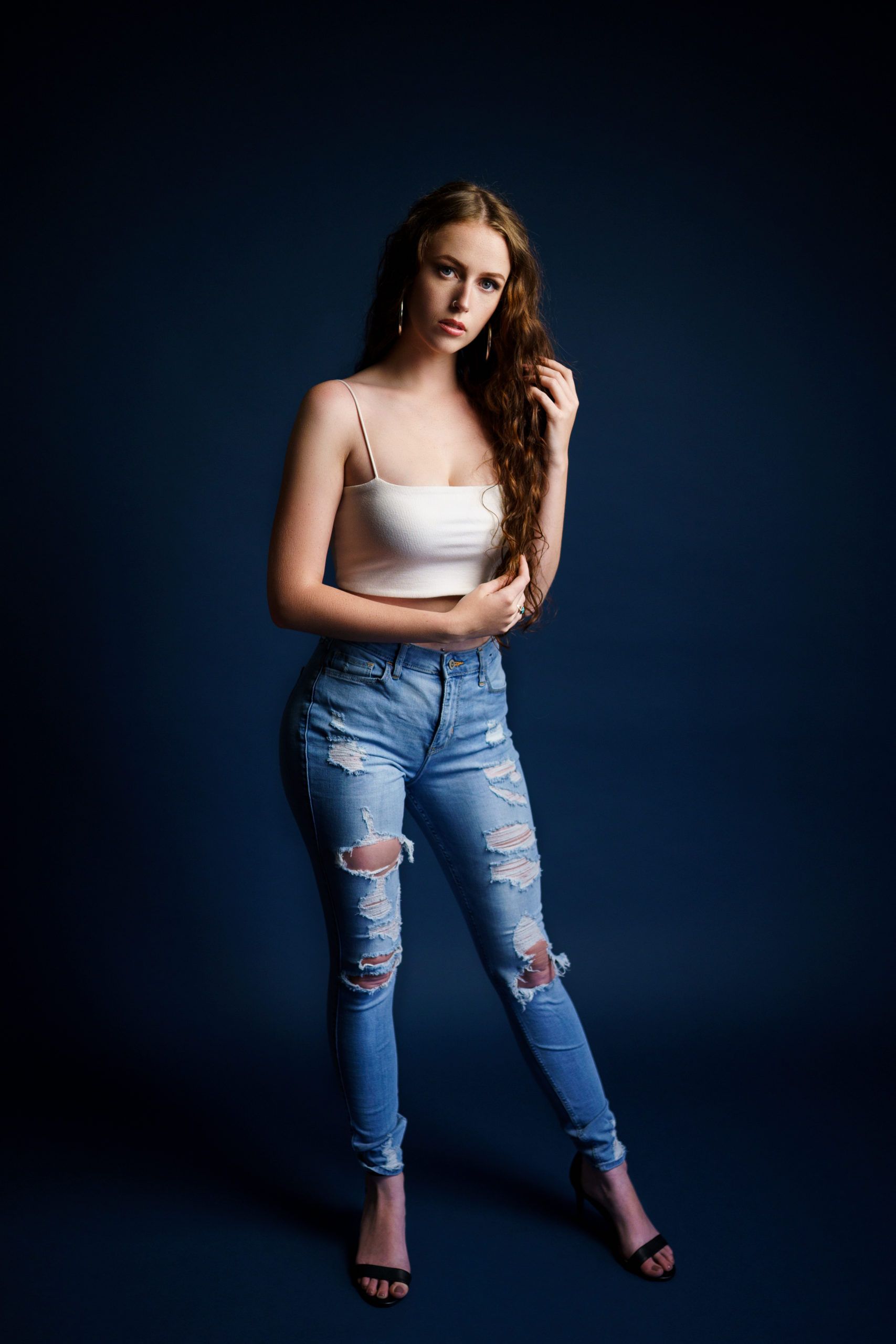 A woman in a white tank top and ripped jeans is standing in front of a dark blue background.
