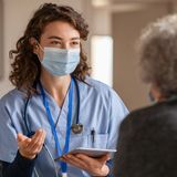 A nurse wearing a mask is talking to an elderly woman.