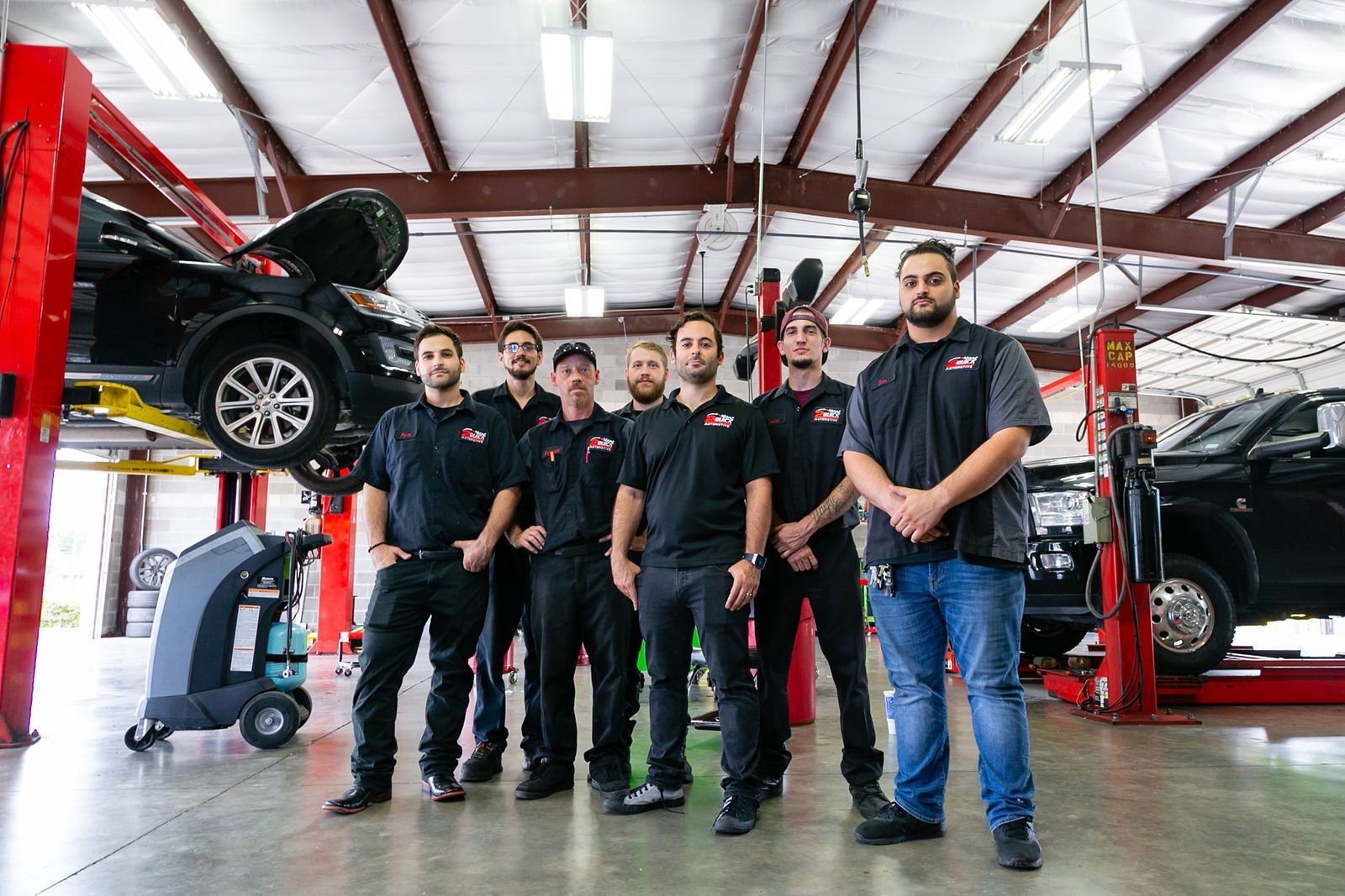 Six mechanics in black uniforms stand in a brightly lit, spacious automotive repair shop with vehicles on lifts. | 2 QuickAuto, Car & Tire Repair | Copperfield