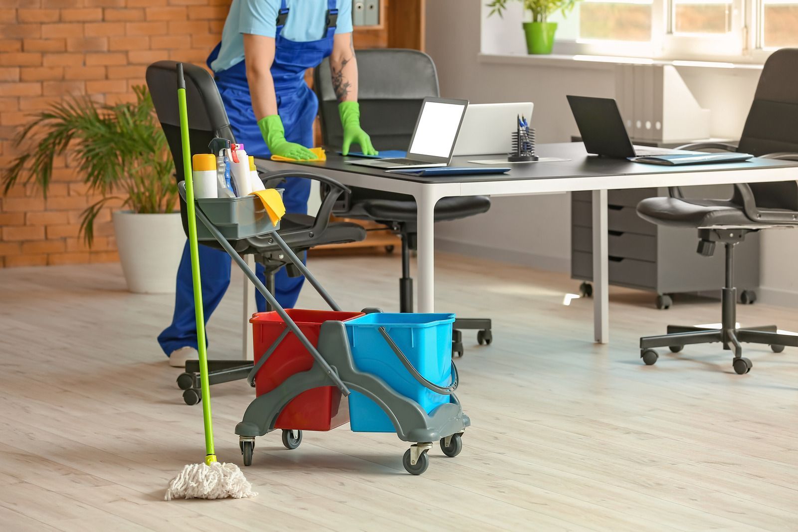 Office cleaner wiping a desk with a laptop. Cleaning supplies in a cart.
