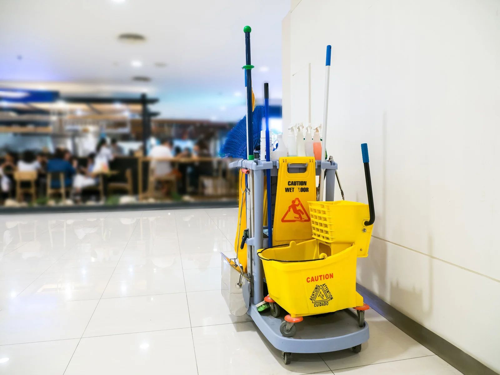 Yellow mop bucket on a white tile floor in a bright hallway, with a blurred cafeteria in the background