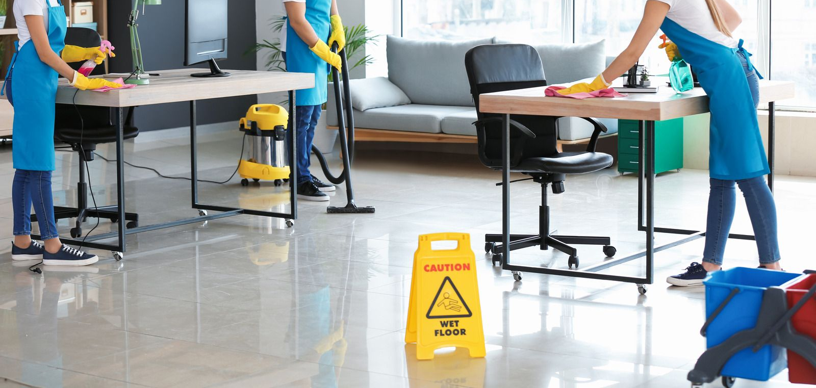 Office cleaning staff mopping a polished floor around desks with a yellow wet floor sign in the center