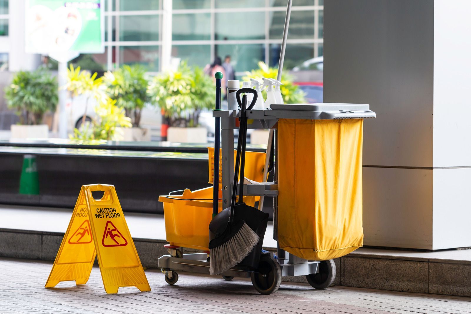 Yellow wet-floor sign beside a janitor cart with mop and trash bag in a building lobby