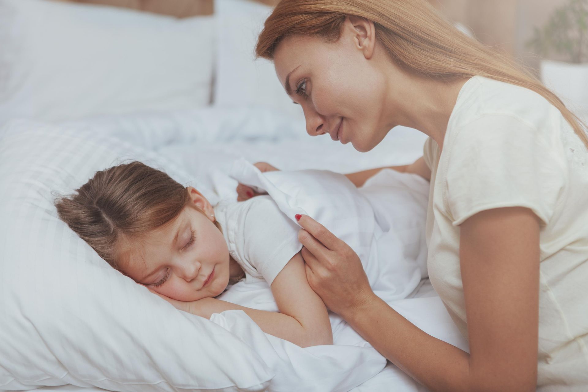 Woman tucks a sleeping child into bed, both are in white clothing and bedding, a bright bedroom.
