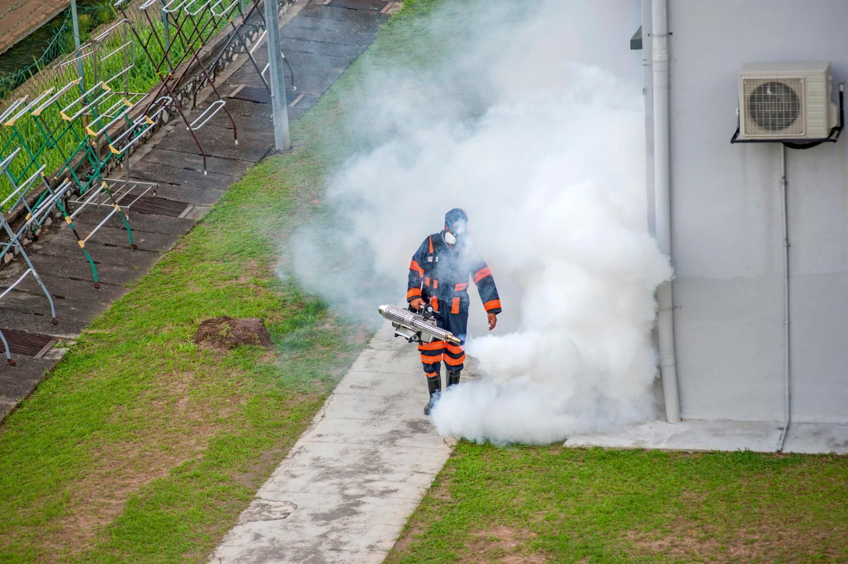 Person in protective suit spraying a white fog outdoors near building with air conditioner.