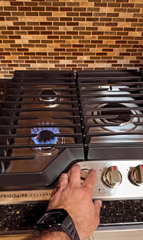Hand adjusting knobs on a stainless gas stove with a blue flame, against a brick backsplash.