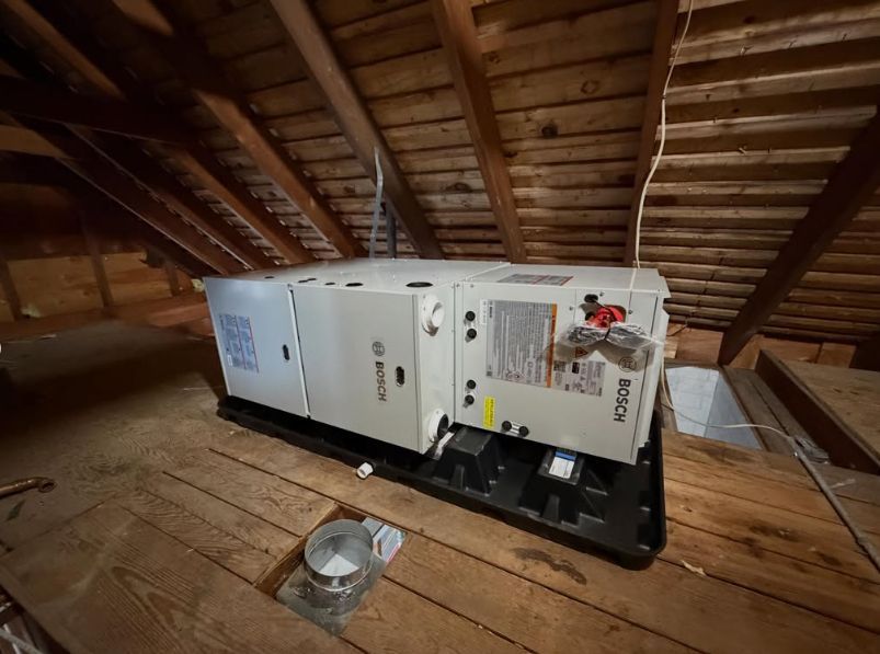 White generator on wooden attic floor beneath sloped rafters, with a bowl in front and a work light beside it